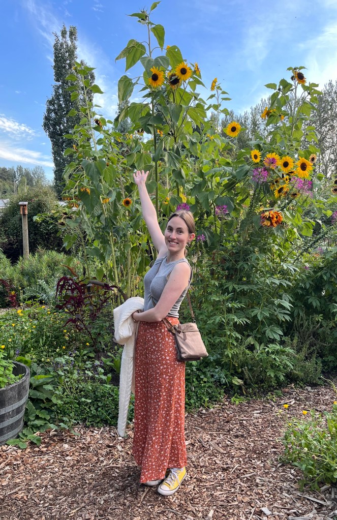 A woman, the website owner, stands with an outstretched arm toward towering sunflowers behind her.